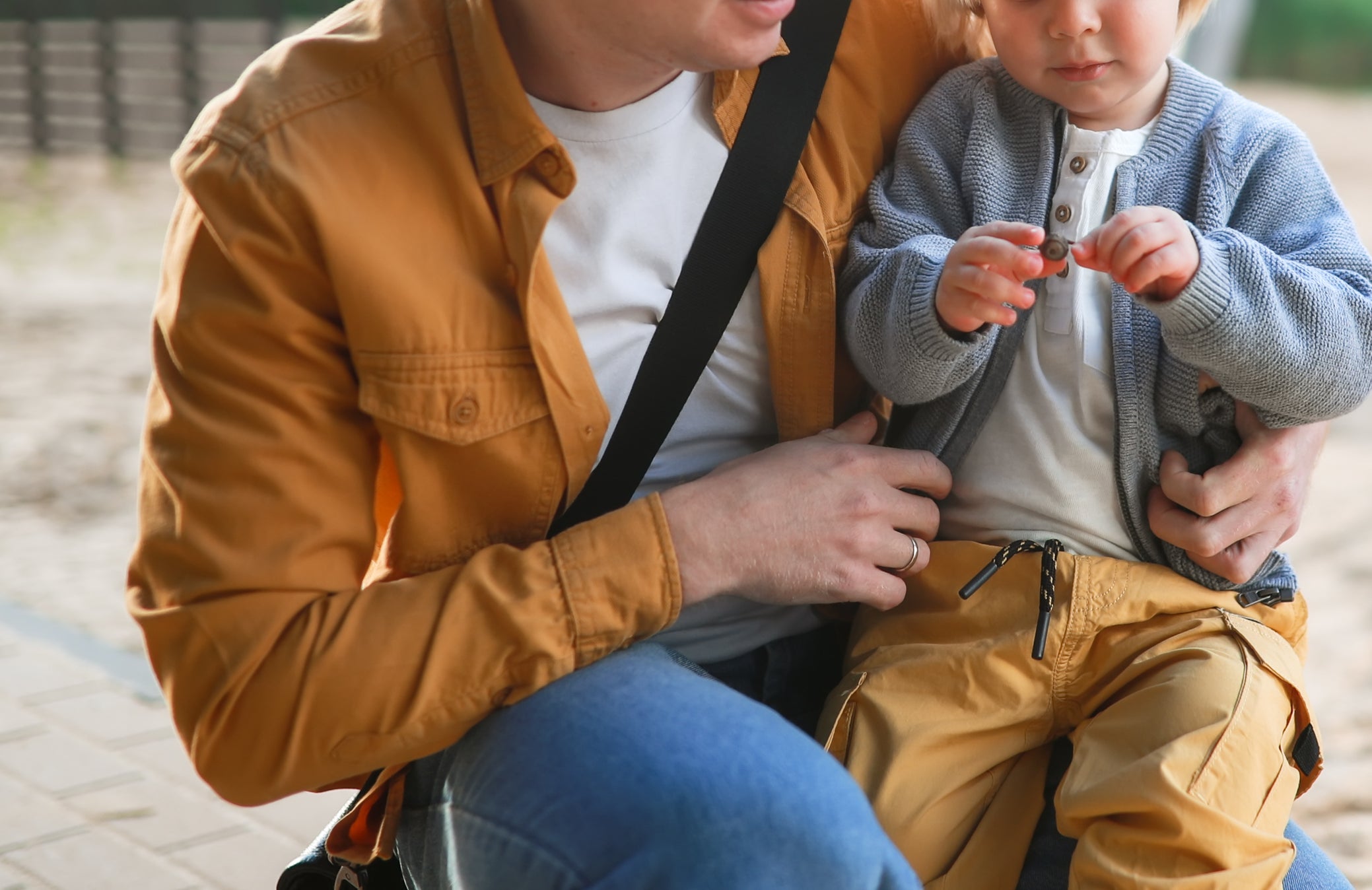 A man with a messenger bag crouches beside a young child at a playground, helping them with what seems to be a small toy or object