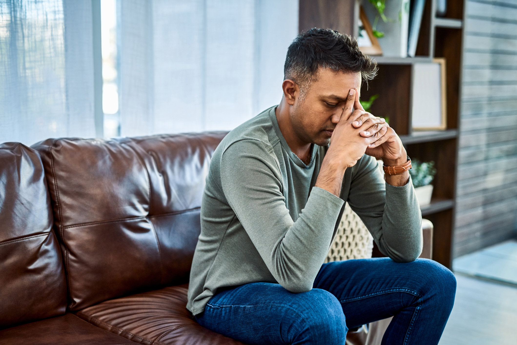 A man in casual clothes sits on a leather couch with his hands clasped, looking contemplative or stressed in a modern home setting