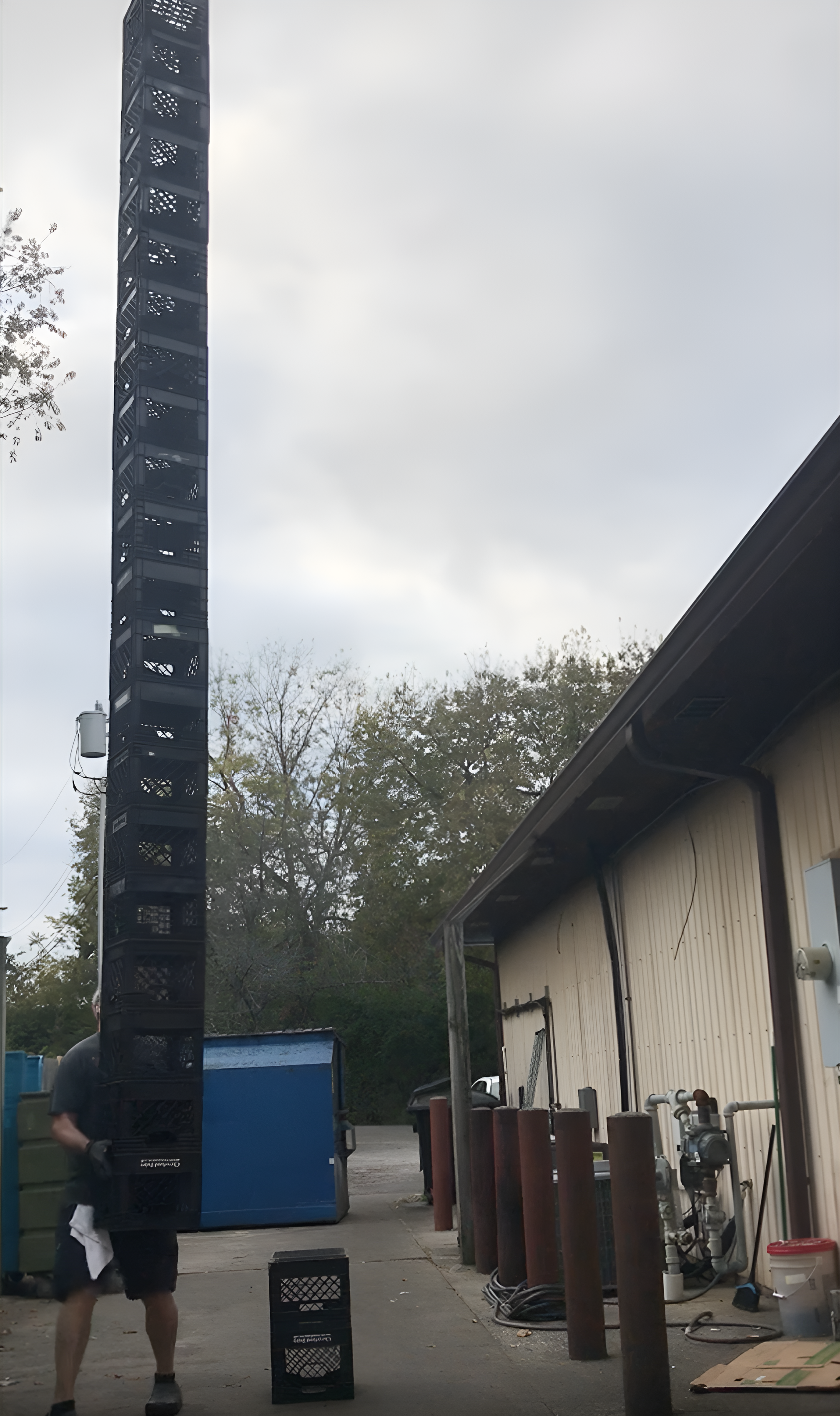 A person balances a tall, precarious stack of milk crates outside a warehouse