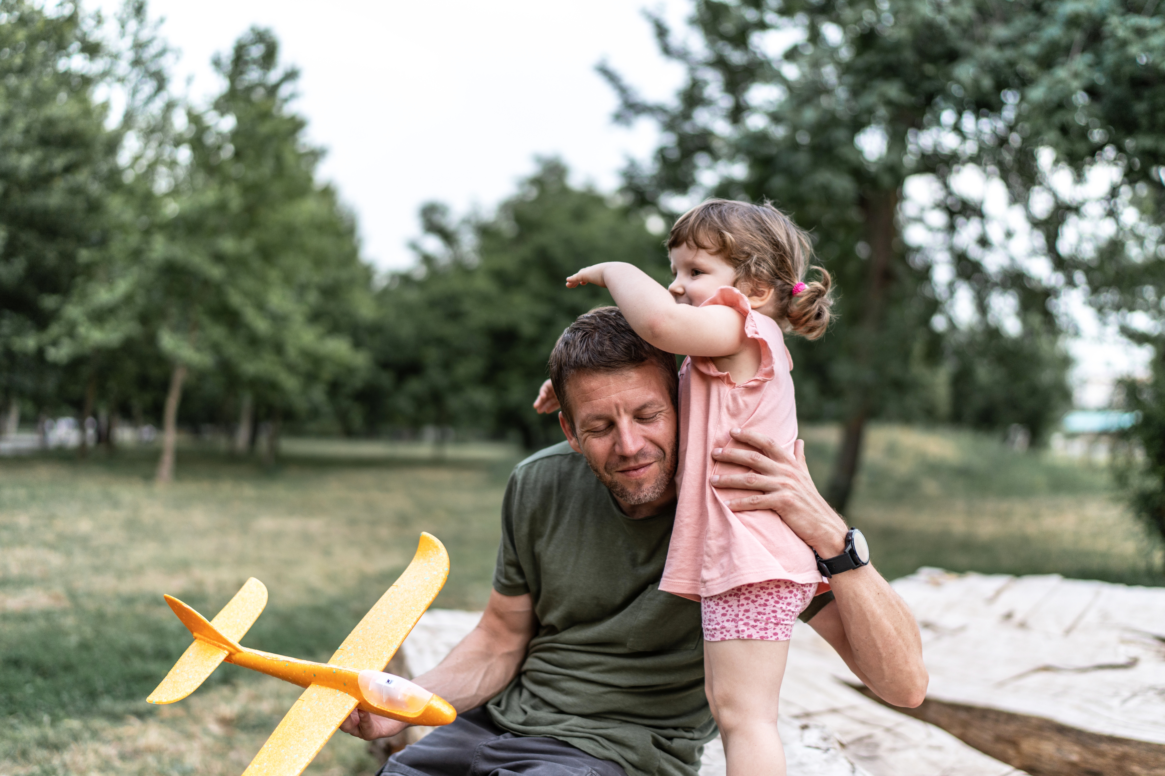 A man sits outdoors holding a toy plane as a young girl in a pink shirt and shorts stands beside him, wiping her face with her arm. They are surrounded by trees