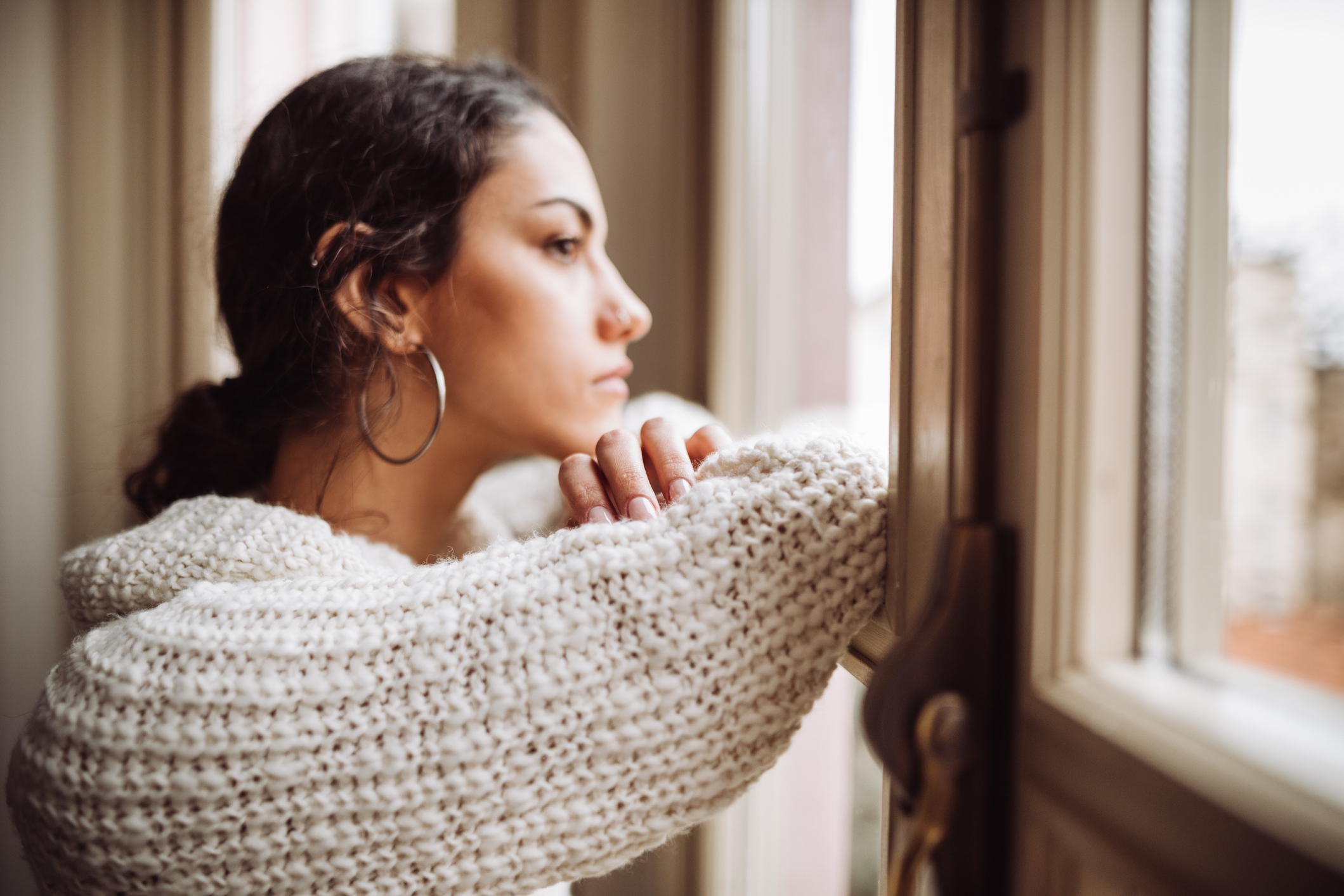 A woman with large hoop earrings and a chunky knit sweater gazes pensively out a window, resting her arms on the windowsill