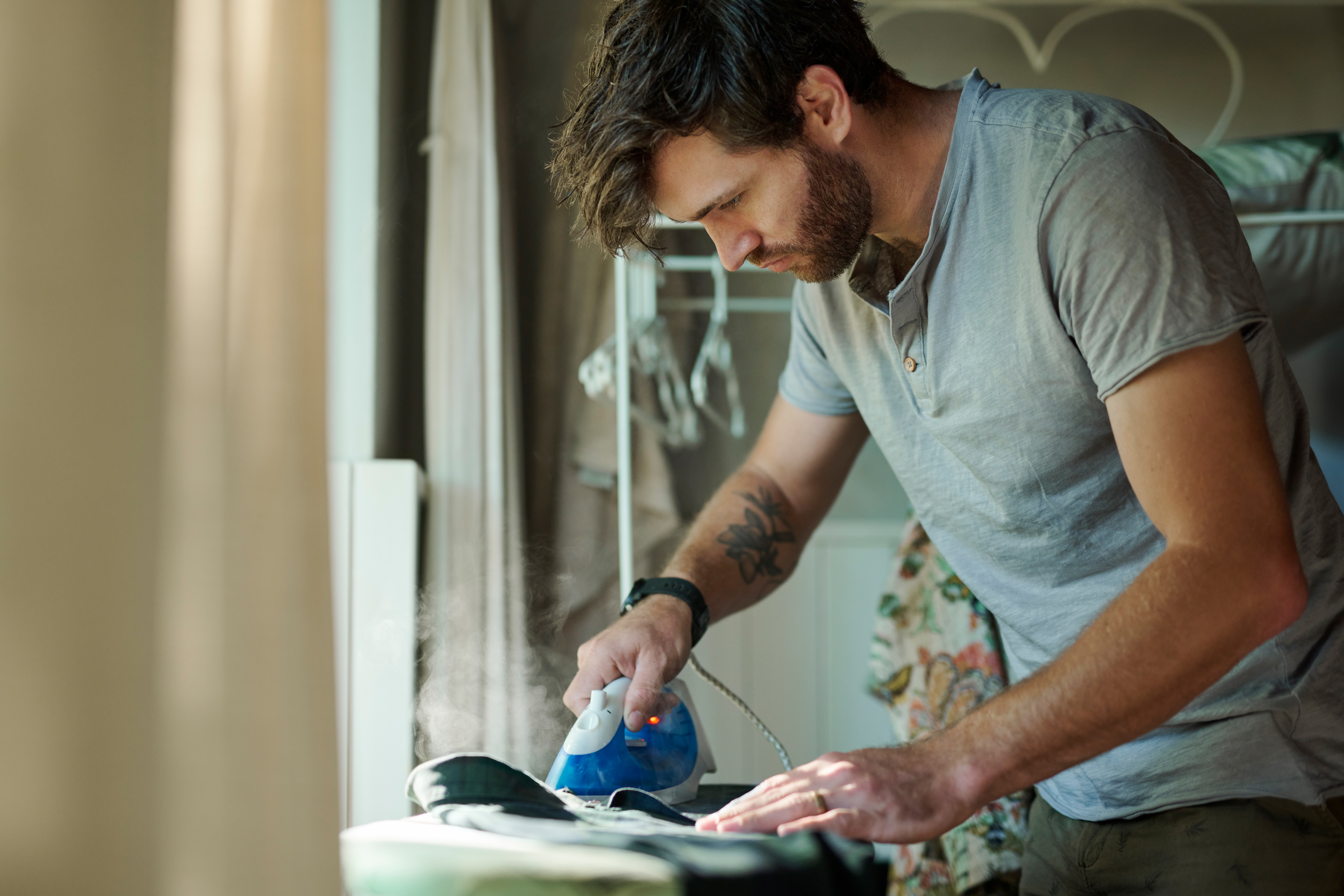 A man with a beard and tattoo on his left arm is ironing clothes on an ironing board in a domestic setting, with steam rising from the iron