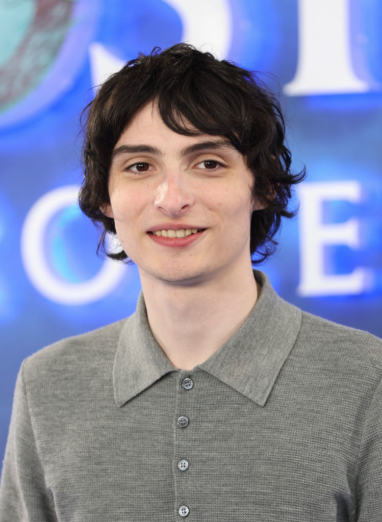 Finn Wolfhard posing in a grey buttoned-up shirt at a celebrity event