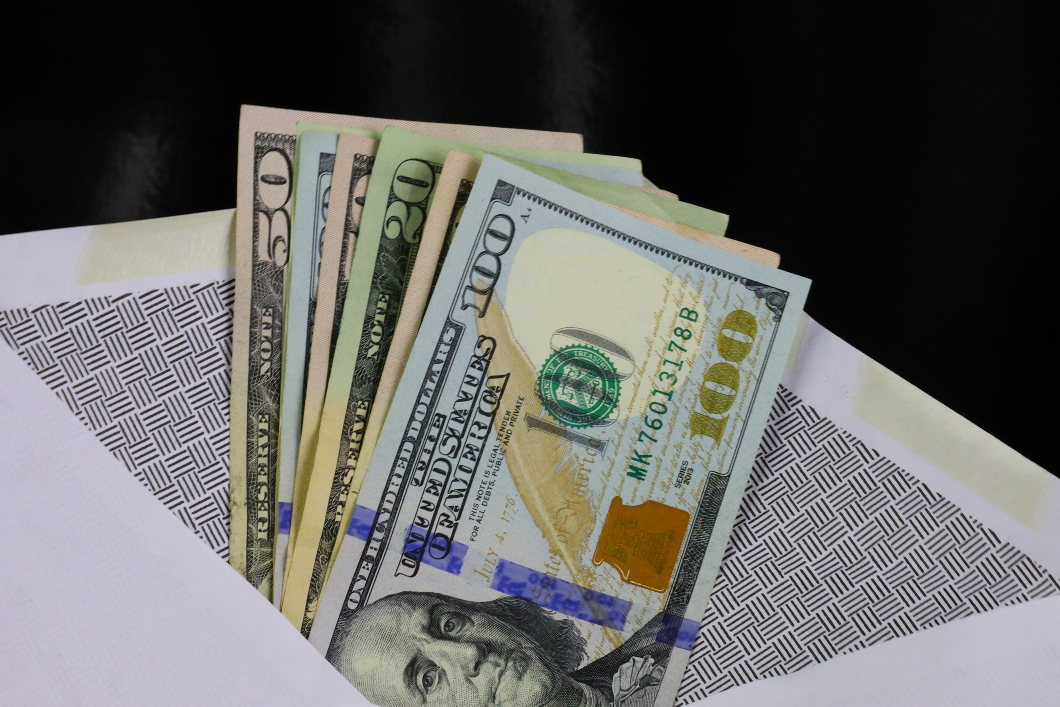 An open envelope revealing multiple US dollar bills, including $100, $50, and $20 denominations, fanned out