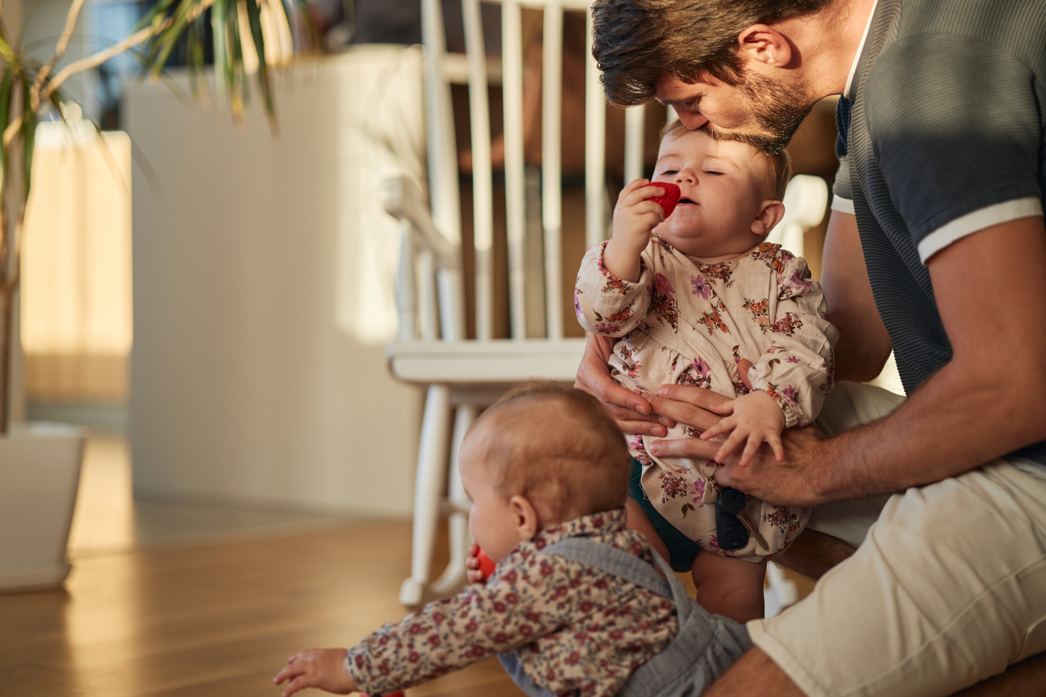 A man kisses a baby on the head while holding them. Another baby in overalls is crawling on the floor