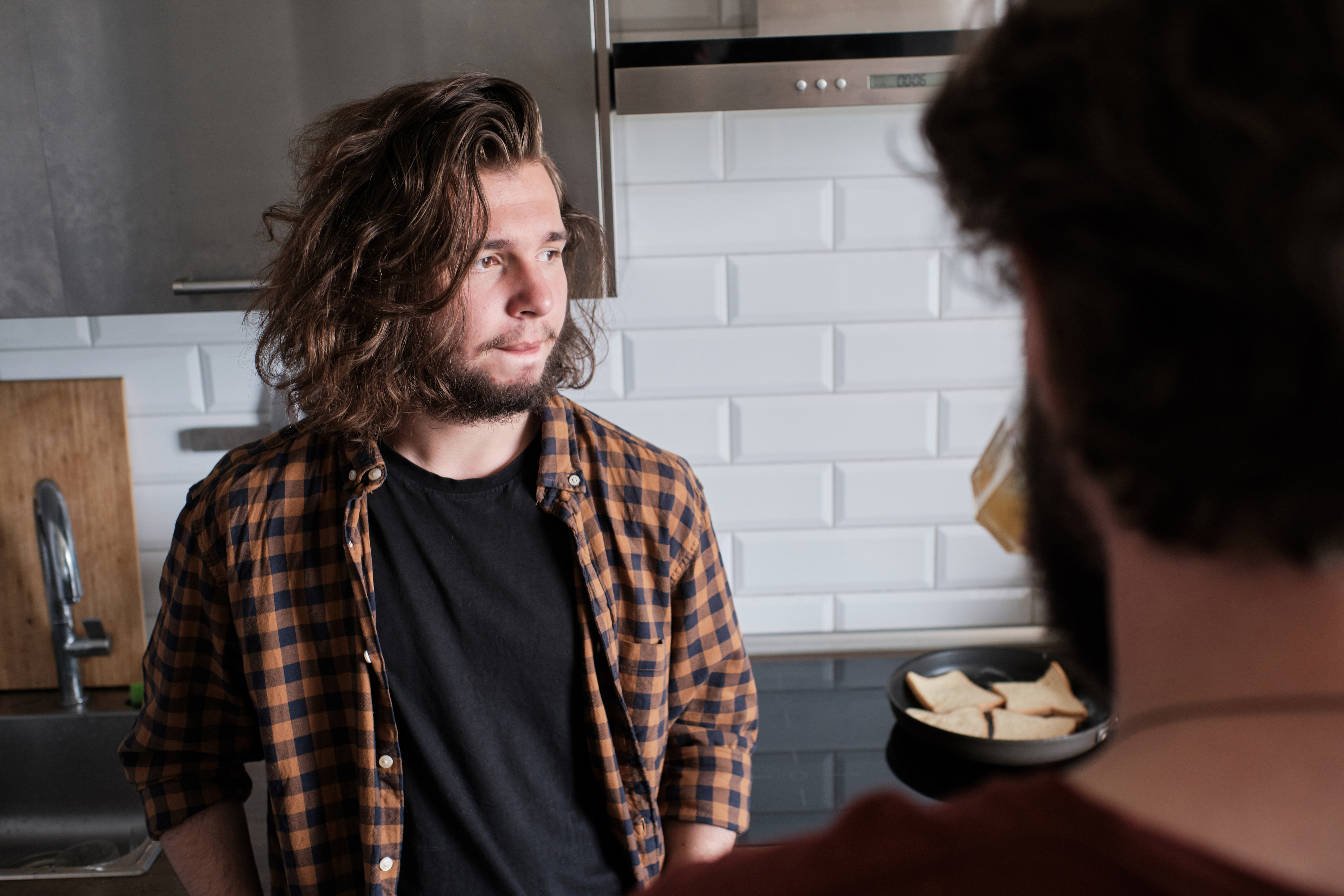 Two men stand in a kitchen talking. One has long hair and a beard, wearing a plaid shirt over a black T-shirt. The other is seen from behind