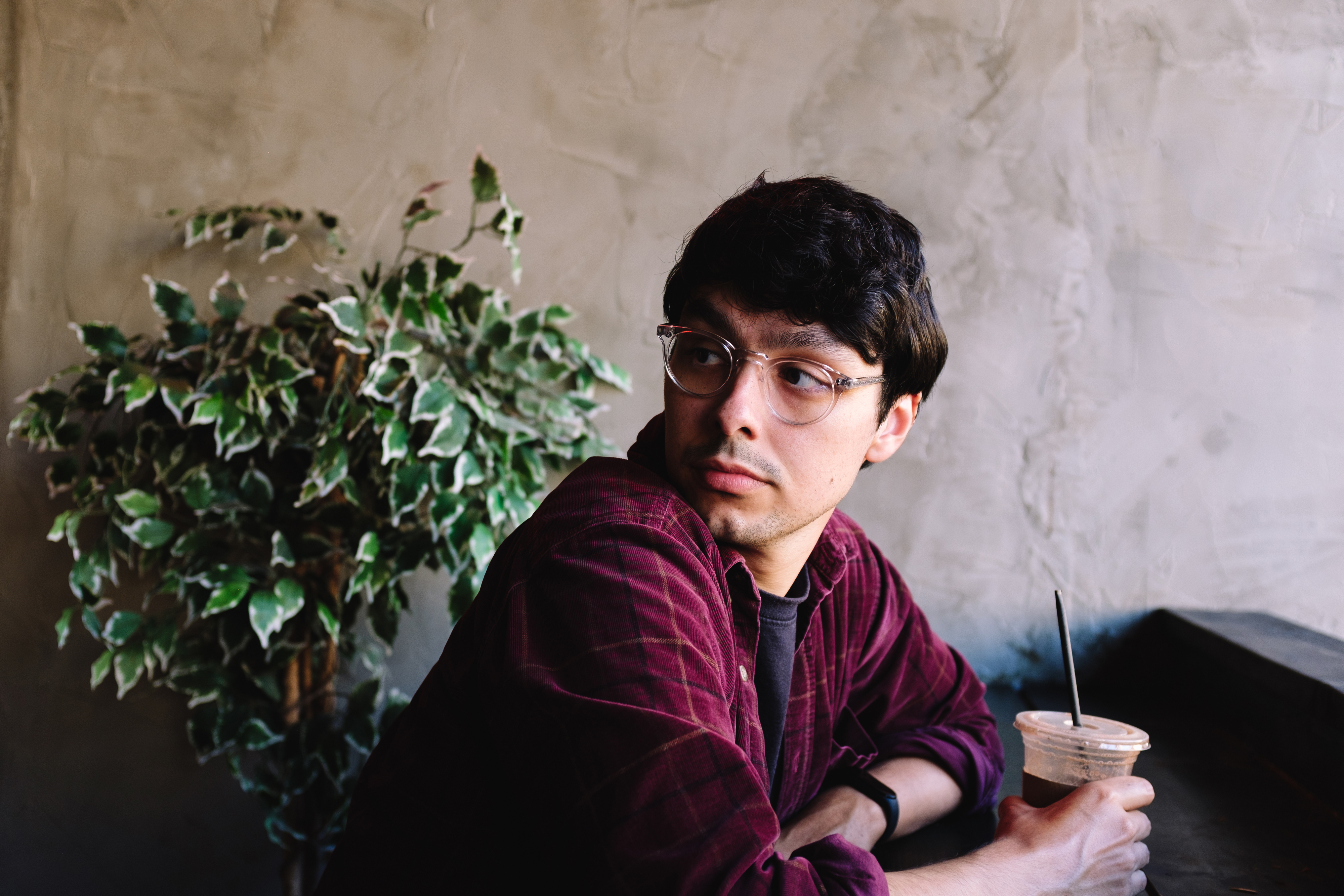 A person with glasses and casual attire sits indoors, holding a drink with a straw, looking off to the side. A plant is in the background