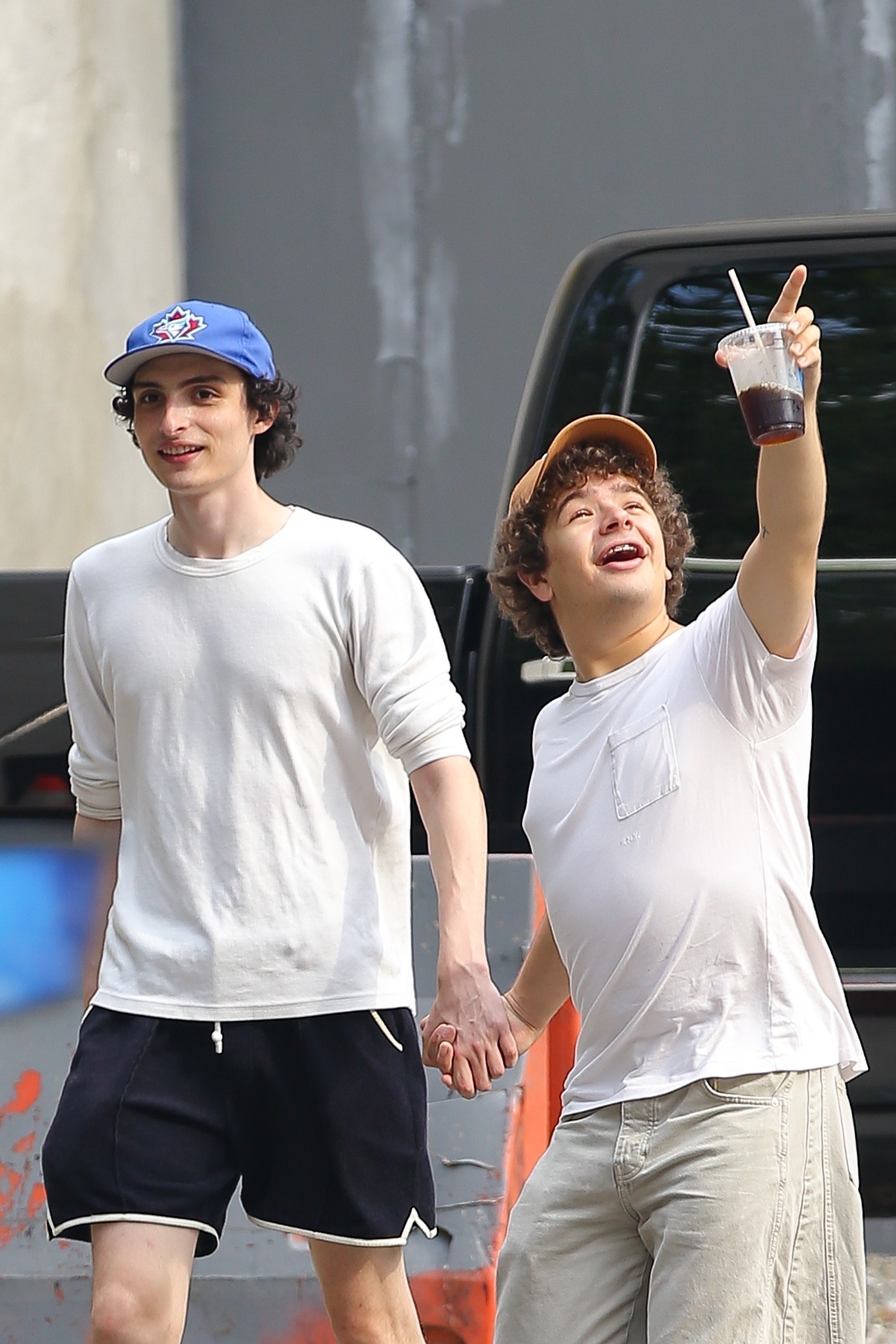 Finn Wolfhard and Gaten Matarazzo hold hands; Finn wears a casual outfit with a baseball cap, and Gaten wears a casual outfit with a cap and holds a drink high