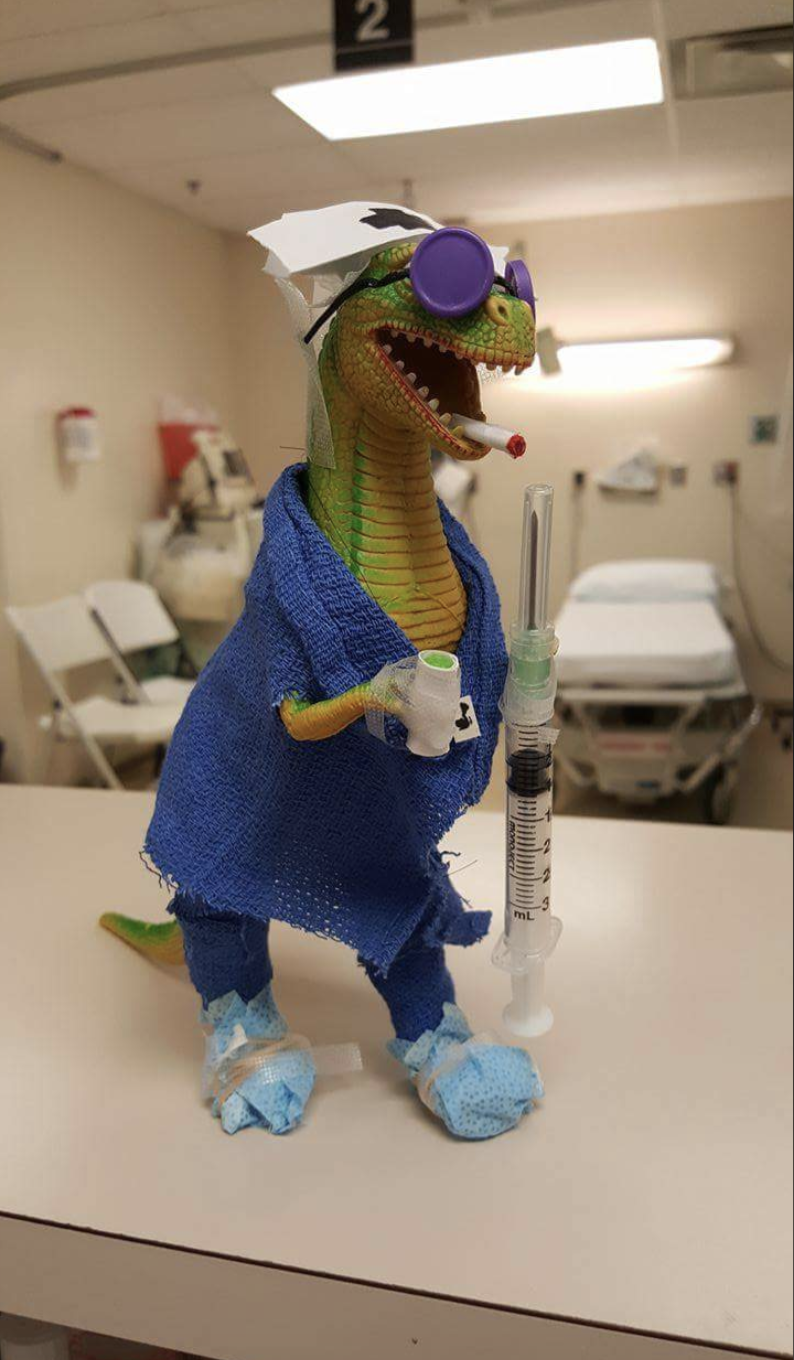 A toy dinosaur is dressed as a patient with a blue gown, a white hat, a medical mask under its chin, sunglasses, and holds a syringe and a medicine cup