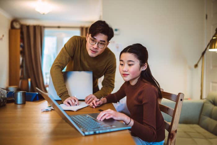 A man and a young girl, both casually dressed, are using a laptop together at a wooden table in a cozy room, engaging in an educational activity