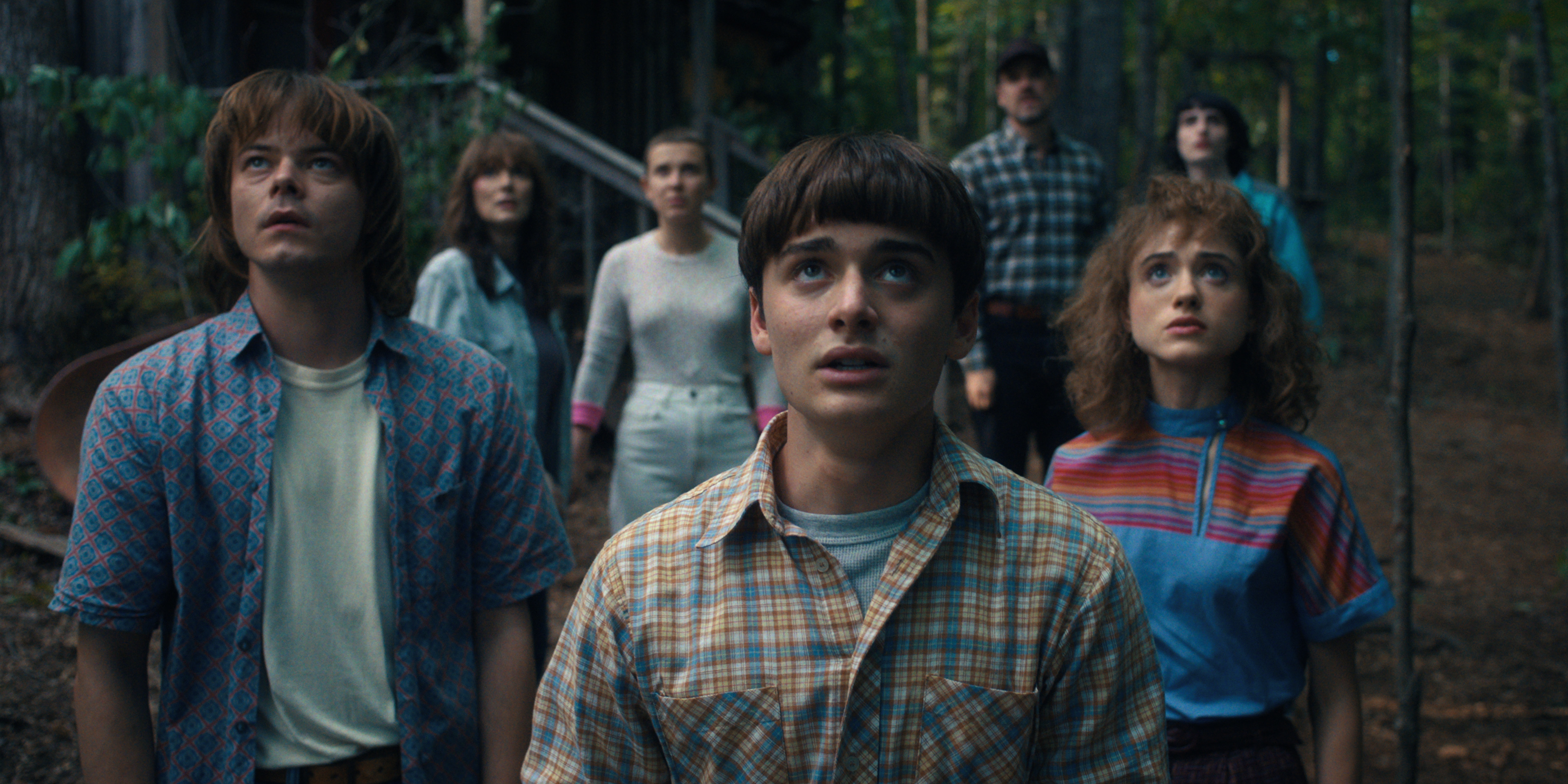 Finn Wolfhard, Charlie Heaton, Millie Bobby Brown, Noah Schnapp, David Harbour, Natalia Dyer, and Maya Hawke look upward in a forest scene from "Stranger Things."
