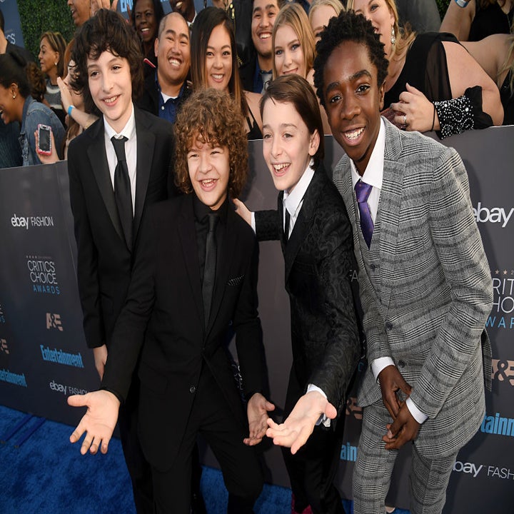 Finn Wolfhard, Gaten Matarazzo, Noah Schnapp, and Caleb McLaughlin in suits pose together at an event in front of a crowd