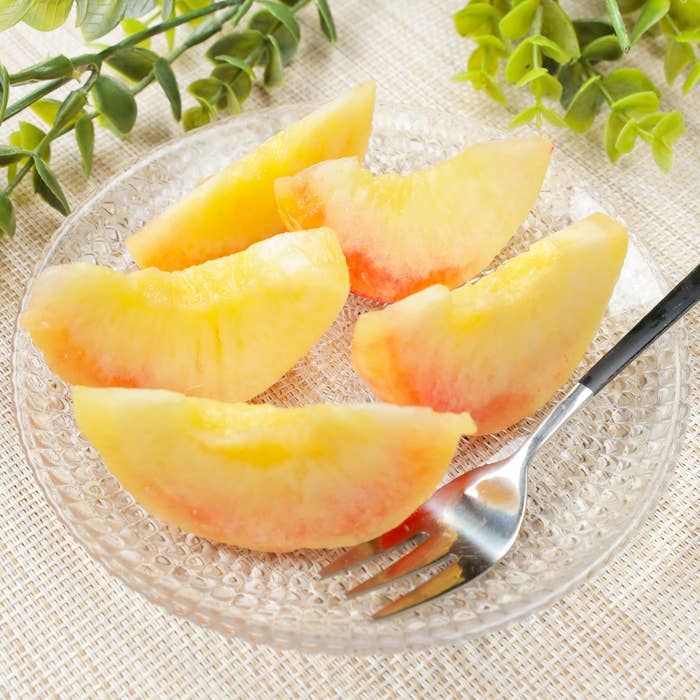 透明な皿に盛り付けられた桃のスライスとフォーク。背景には緑の植物が置かれている。