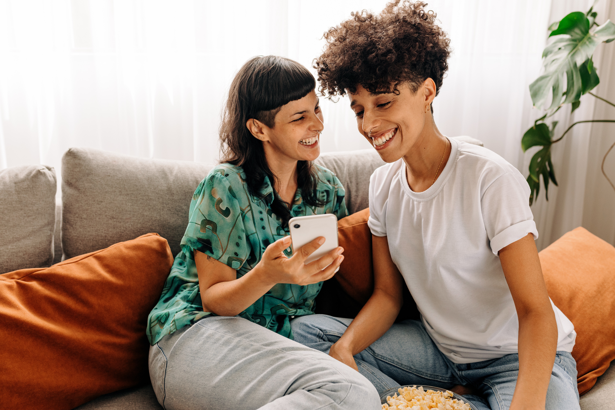 Two people sit on a couch smiling while one shows something on a phone screen to the other