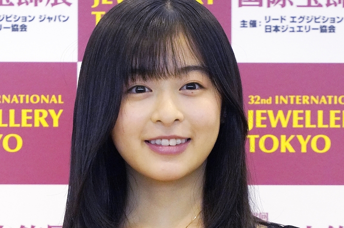 女性（名前不明）が国際宝飾展のイベントで黒いドレスを着て笑顔を見せています。背景にはイベントの名前が表示されています。
