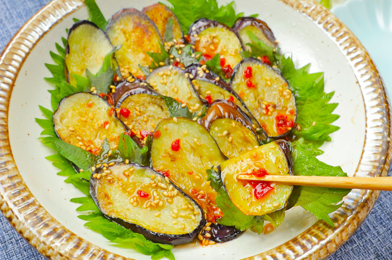 お皿に盛られた茄子の料理。ゴマや赤いトッピングが施され、箸で一切れが持ち上げられている。緑の葉が添えられている。