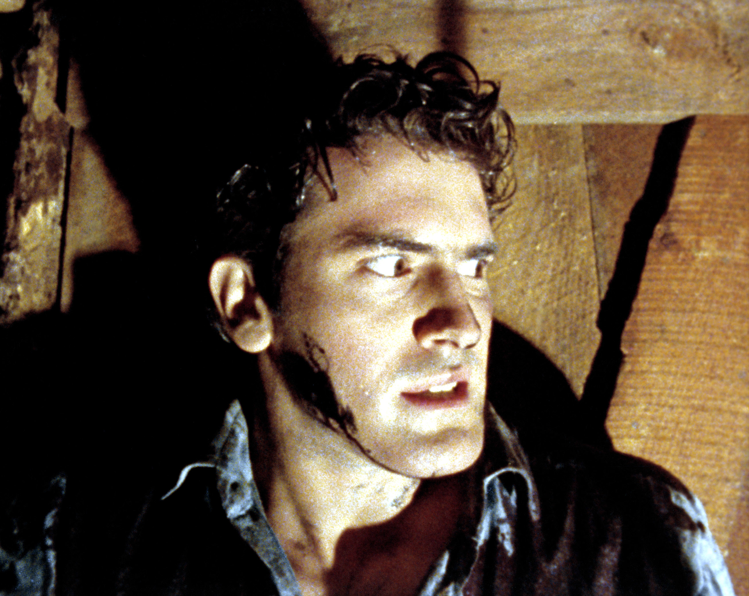 Bruce Campbell, in a distressed state with dirt and smudges on his face, leans against a wooden wall, appearing alarmed in a scene from &quot;The Evil Dead&quot;