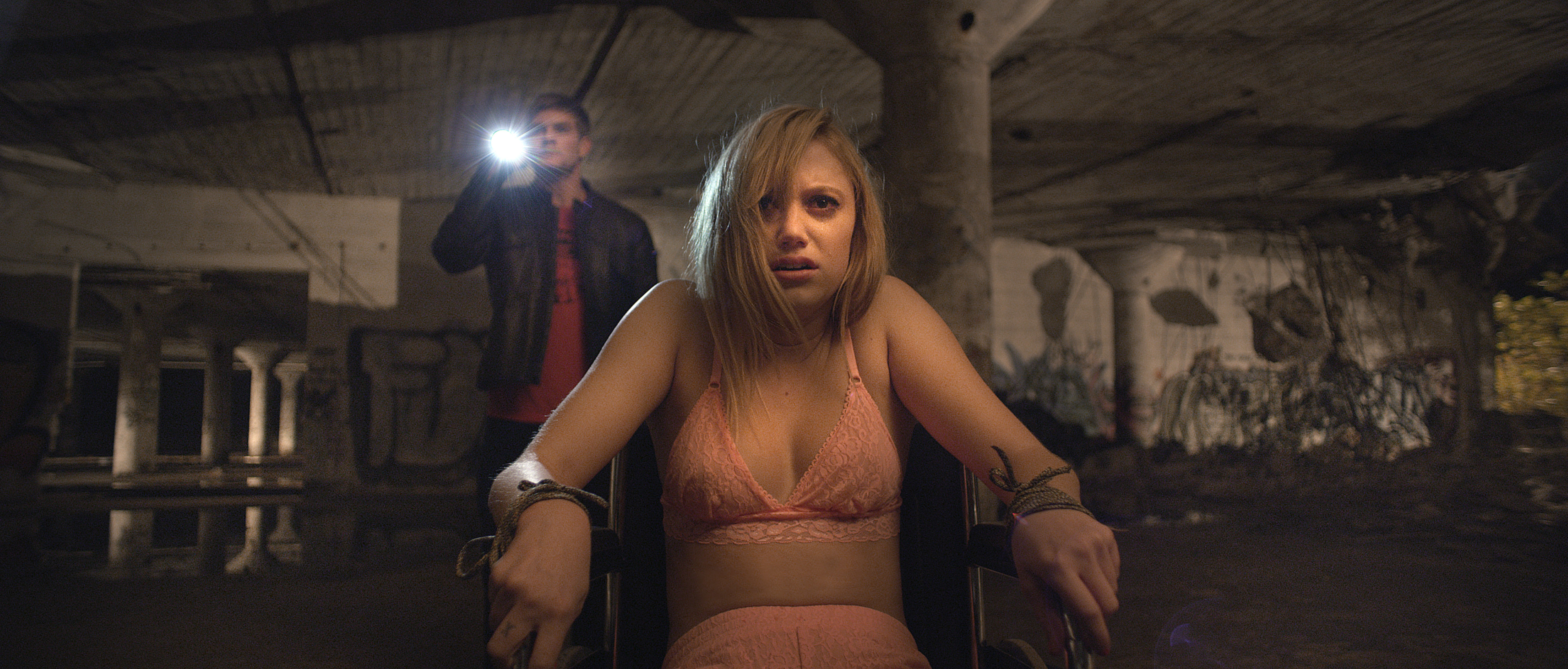 Maika Monroe sitting in a chair with her hands tied and a man holds a flashlight behind her in a scene from &quot;It Follows&quot;