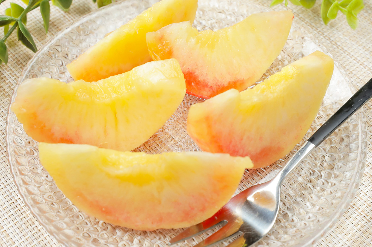 透明の皿に盛られた4切れの桃とフォークが見える。葉っぱが周りに配置されている。