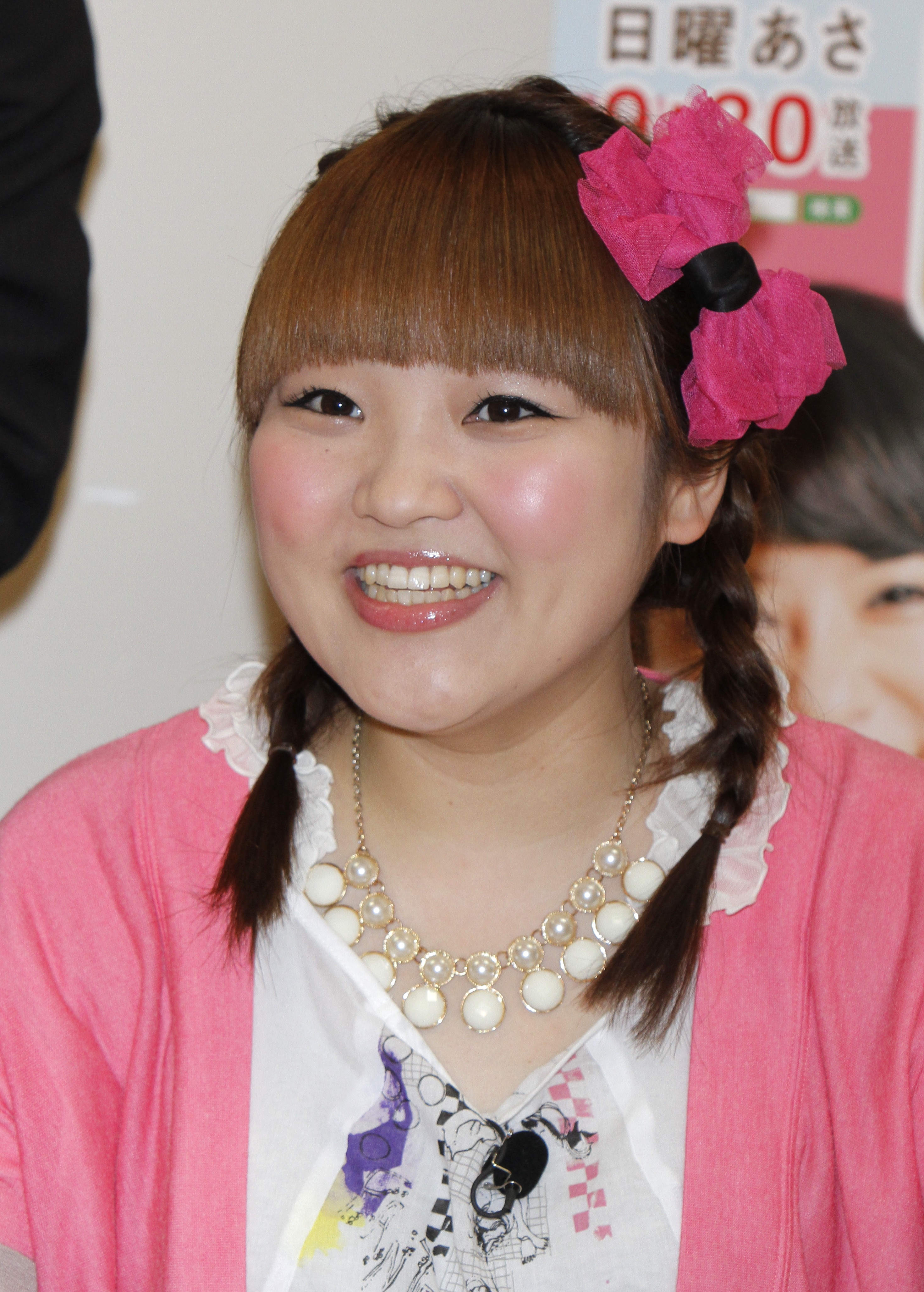 女性がカメラに向かって笑顔を見せています。彼女はピンクのヘアアクセサリーを付け、カジュアルなスタイルの服を着ています。