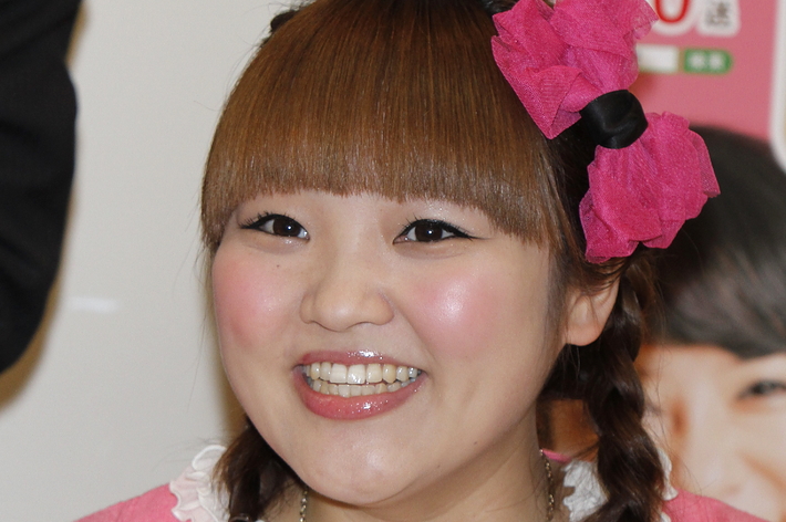 女性がカメラに向かって笑顔を見せています。彼女はピンクのヘアアクセサリーを付け、カジュアルなスタイルの服を着ています。