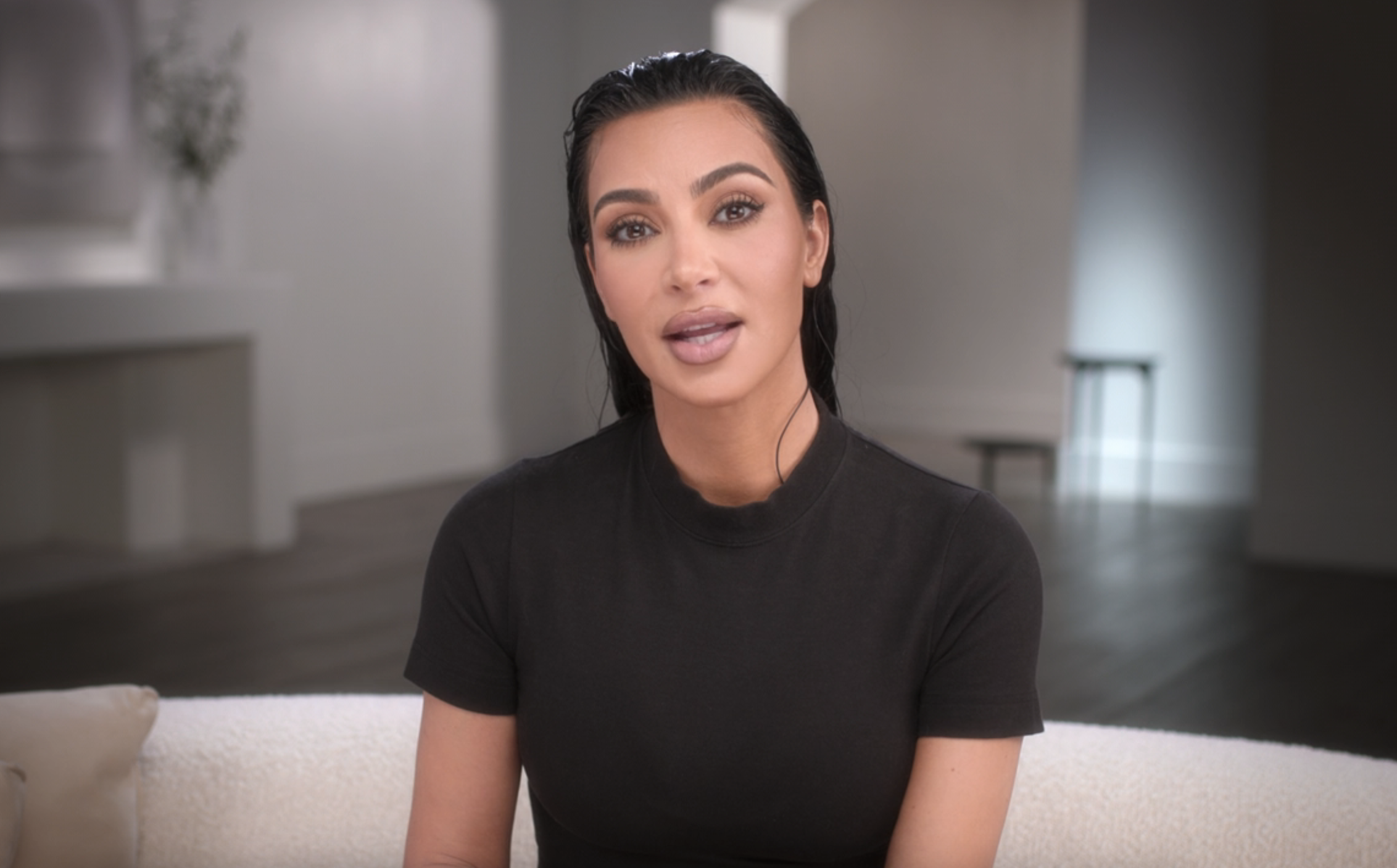 Kim Kardashian sitting on a couch, wearing a simple black top, with a minimalist background behind her