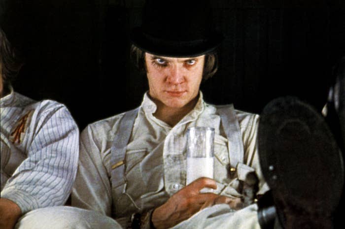 A man dressed in white with suspenders, wearing a black bowler hat, sits and holds a glass of milk, with a serious expression on his face