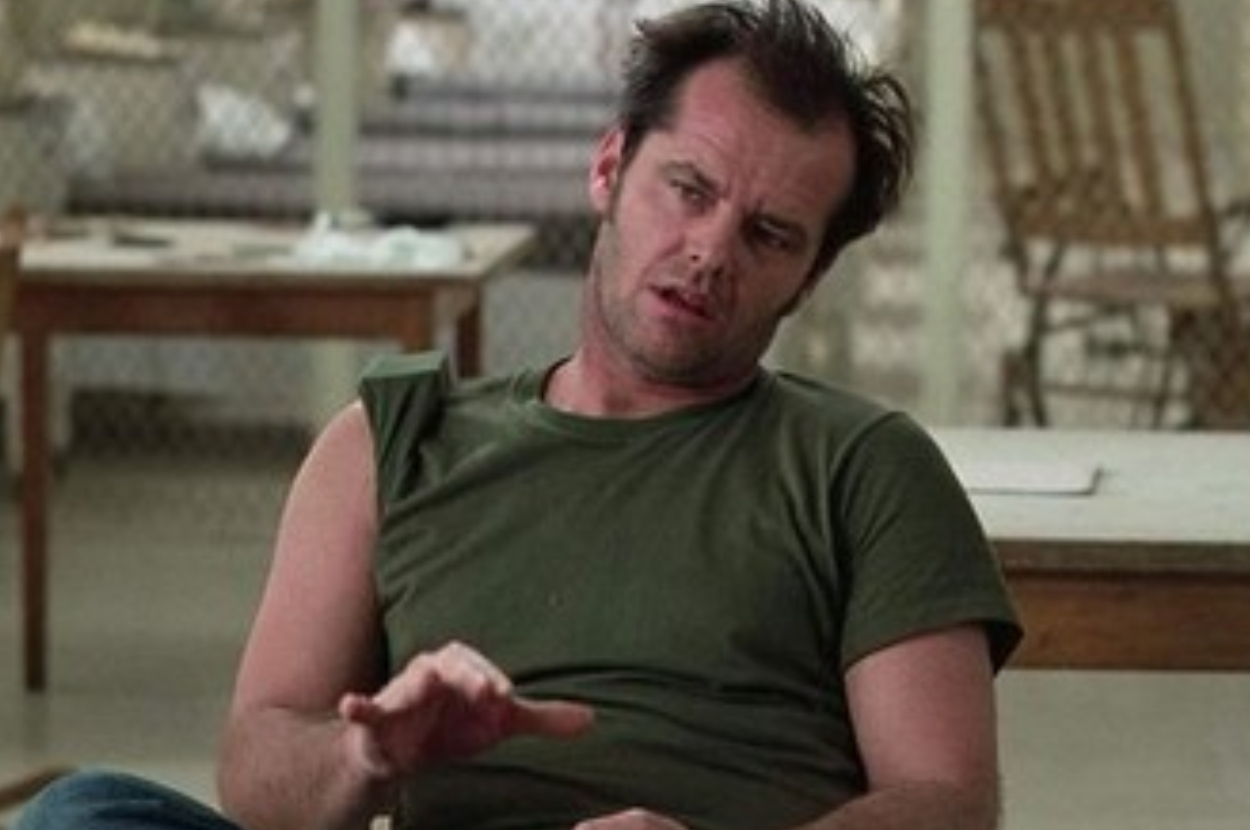 A man with disheveled hair, wearing a torn green T-shirt, is seated and talking in a room with a table and chairs in the background