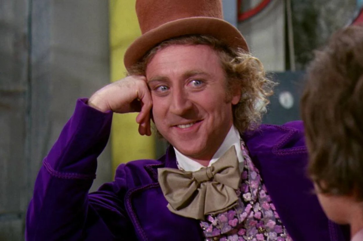 Gene Wilder as Willy Wonka in "Willy Wonka & the Chocolate Factory," wearing a large bow tie, floral shirt, and purple coat, rests his head on his hand