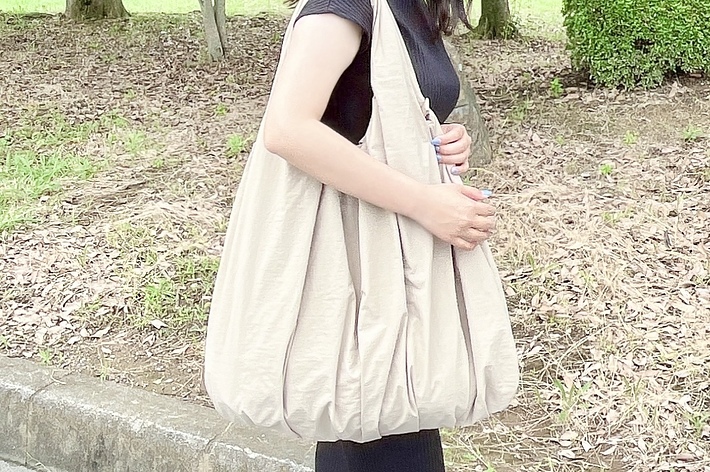 女性が公園の歩道を歩いています。肩に大きなベージュのバッグをかけ、黒いドレスを着ています。