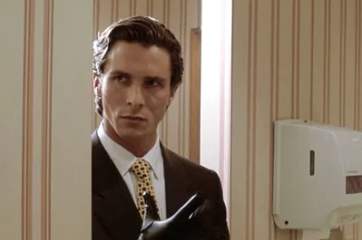 Christian Bale as Patrick Bateman, dressed in a suit with a patterned tie, glancing from behind a wall in a scene from the film "American Psycho."