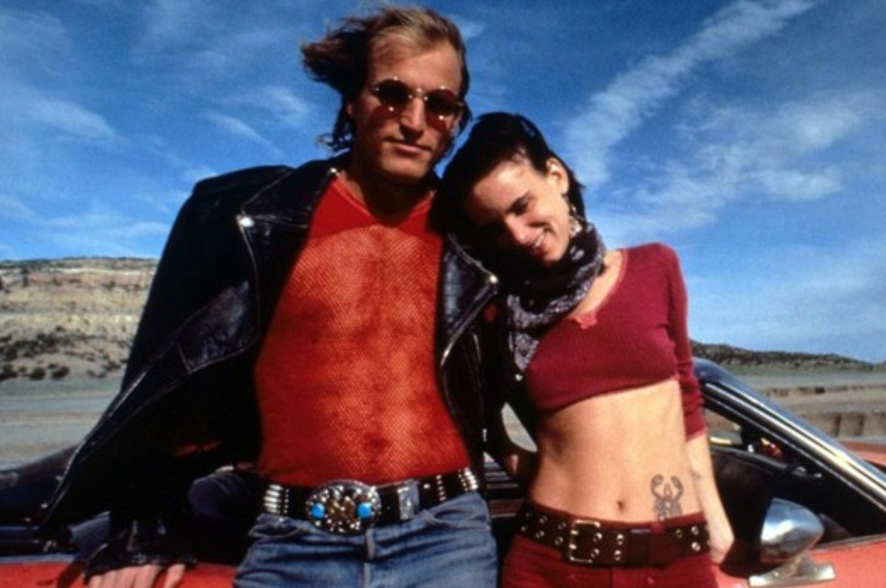 Woody Harrelson and Juliette Lewis standing in front of a red car, Harrelson in a leather jacket and fishnet shirt, and Lewis in a crop top with a visible tattoo