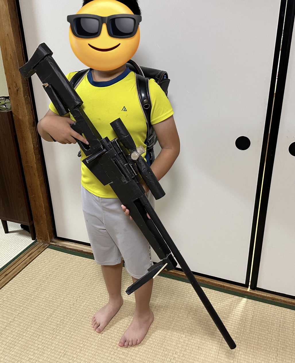 男の子が手作りの大きなおもちゃの銃を持ち、サングラスの絵文字で顔が隠されています。