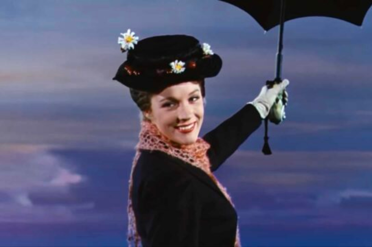 Julie Andrews as Mary Poppins smiles while holding an umbrella against a sky backdrop. She wears a black hat adorned with flowers, a red scarf, and gloves