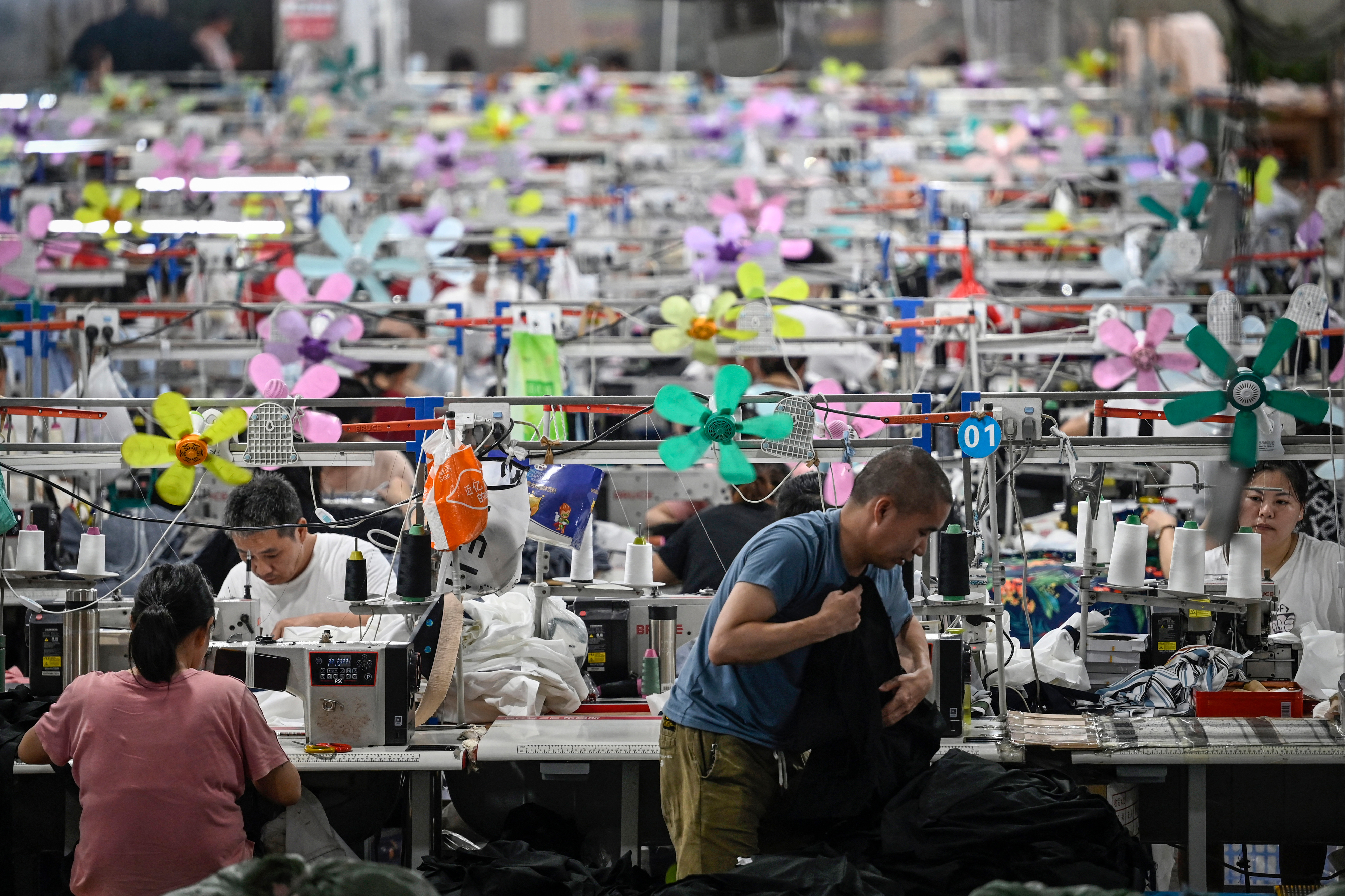 People at work stations in a large garment factory. Sewing machines are surrounded by colorful small fans and various textiles