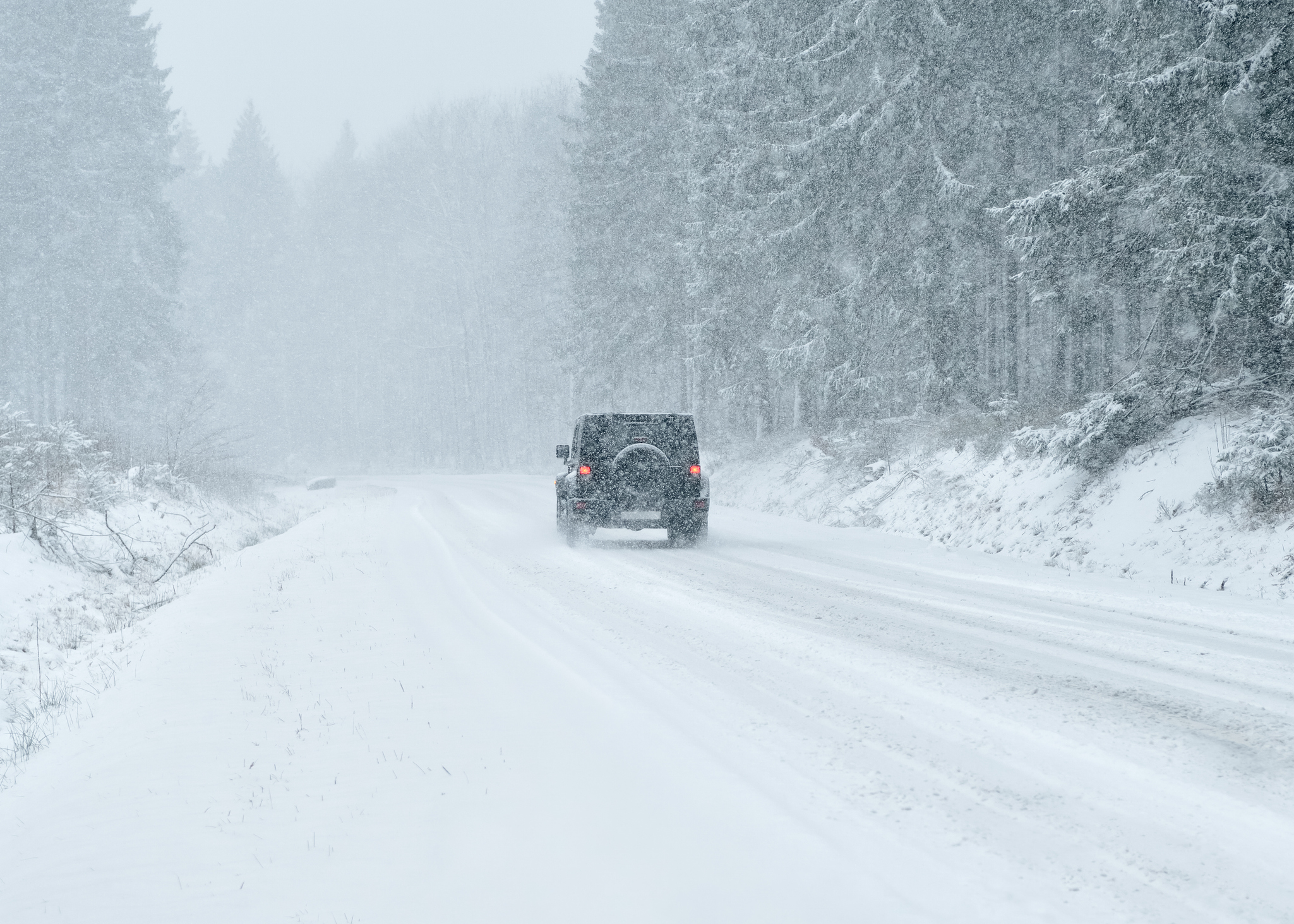 A car is driving on a snowy, tree-lined road during heavy snowfall, surrounded by frosted trees and snow-covered ground