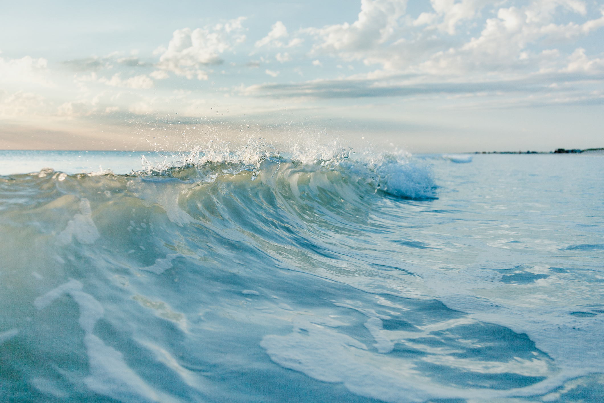 Ocean wave crashing gently under a partly cloudy sky with the horizon in the background