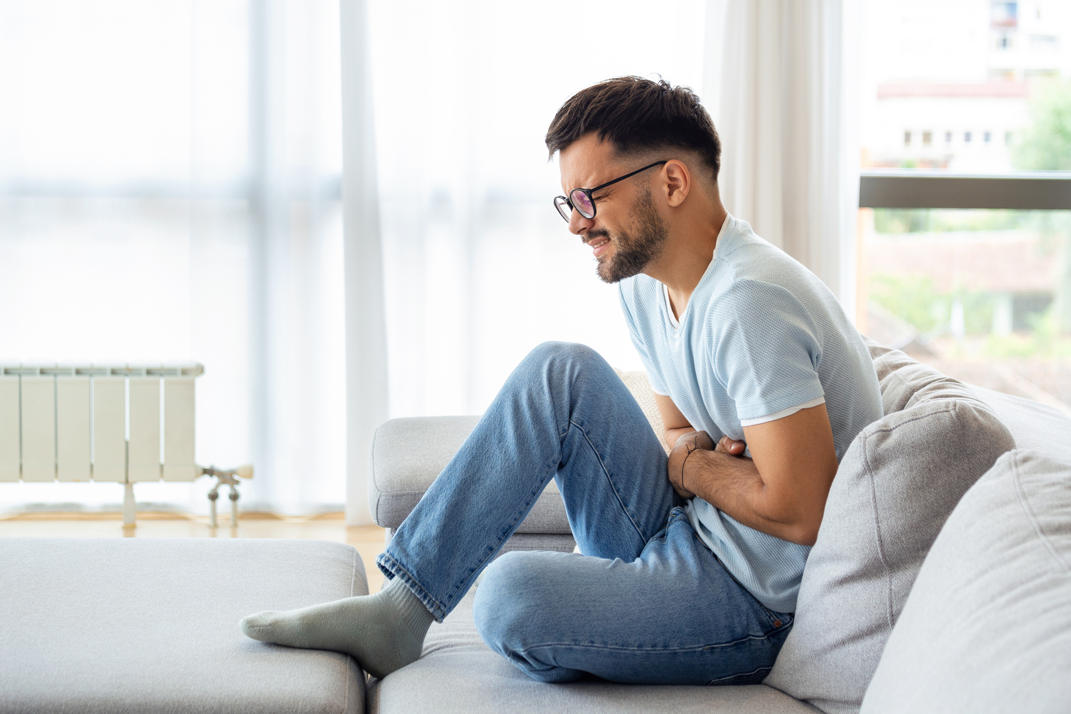 A man with short hair and glasses sits on a couch, holding his stomach in discomfort while wearing a light shirt and jeans