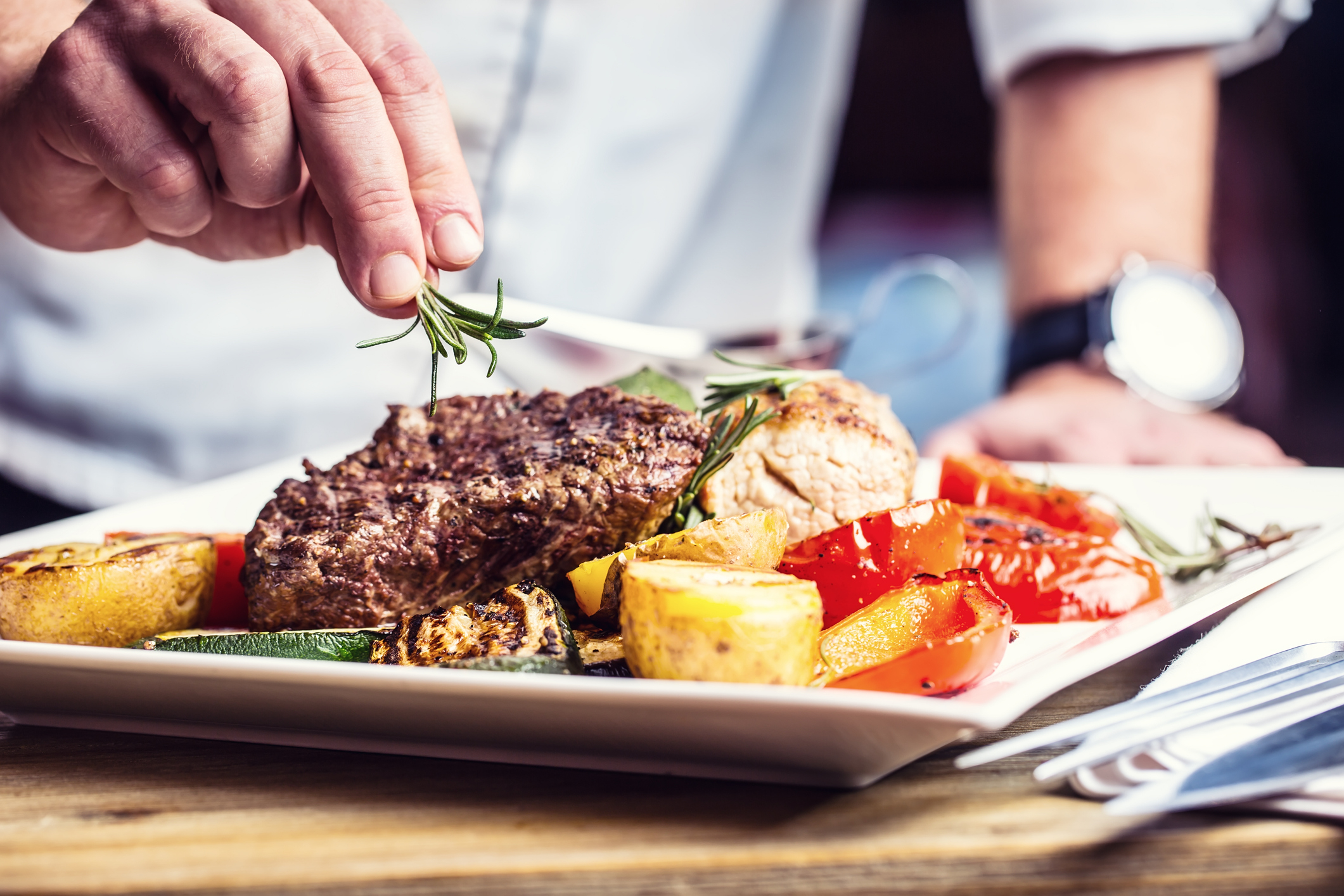 A chef garnishes a plate of steak and roasted vegetables with a sprig of rosemary
