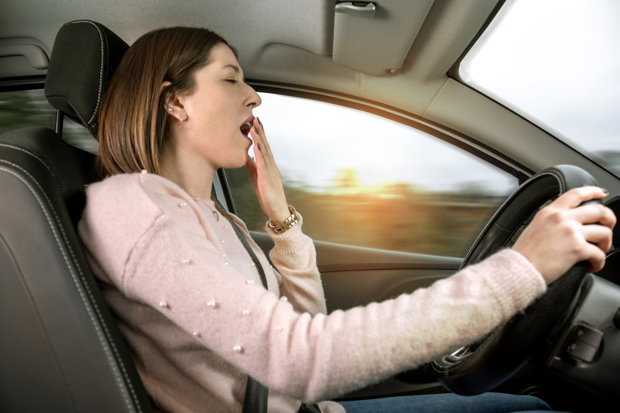 A woman yawns while driving a car, covering her mouth with one hand