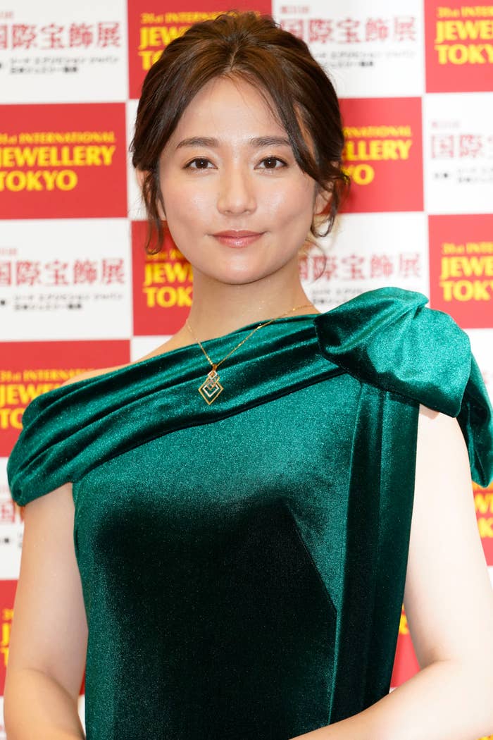 女優が東京国際宝飾展のレッドカーペットイベントでエレガントなドレスを着ています。背景にイベントのロゴがあります。