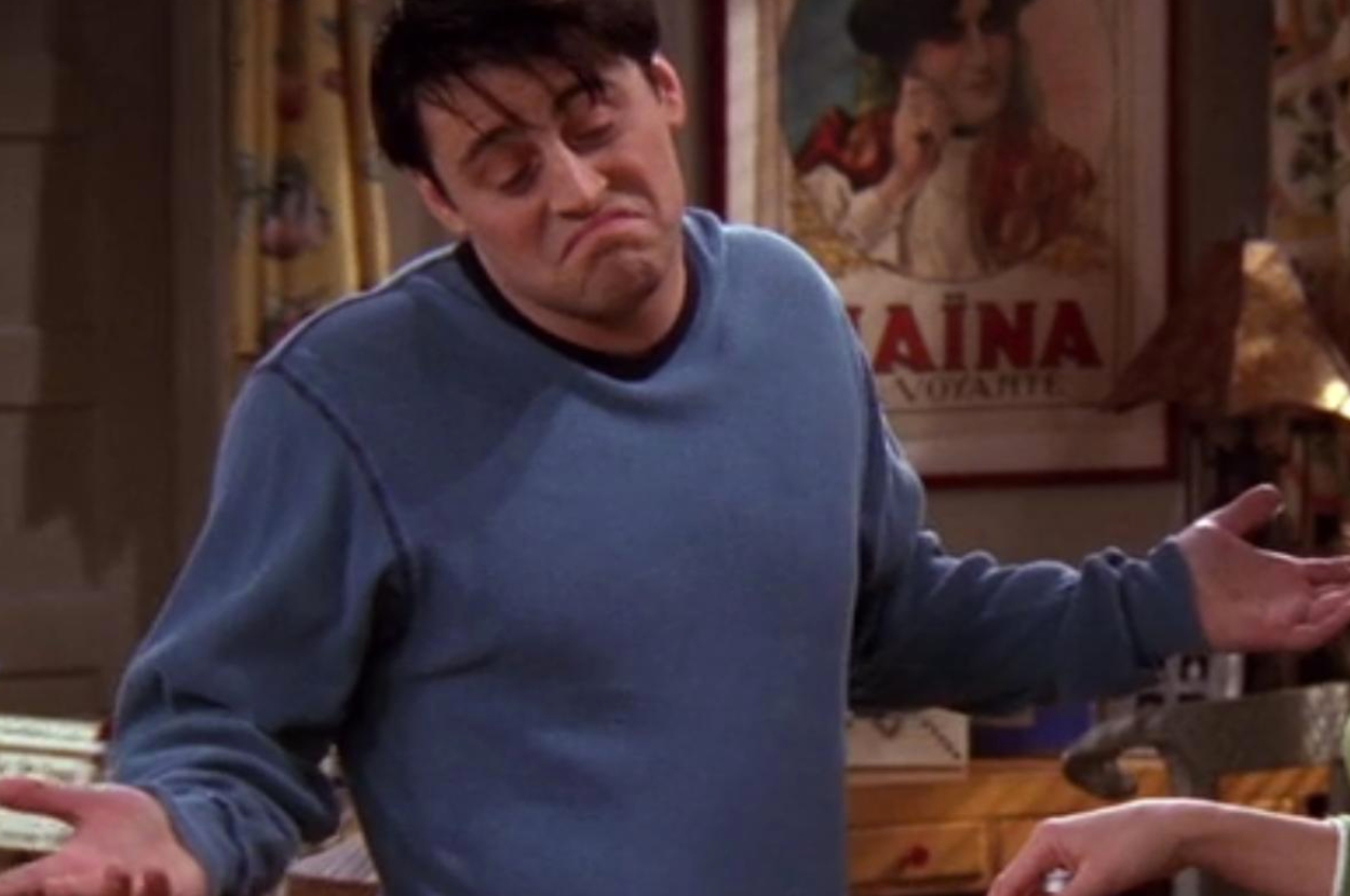 Man wearing a blue long-sleeve shirt shrugs with a nonchalant expression, standing indoors with posters in the background