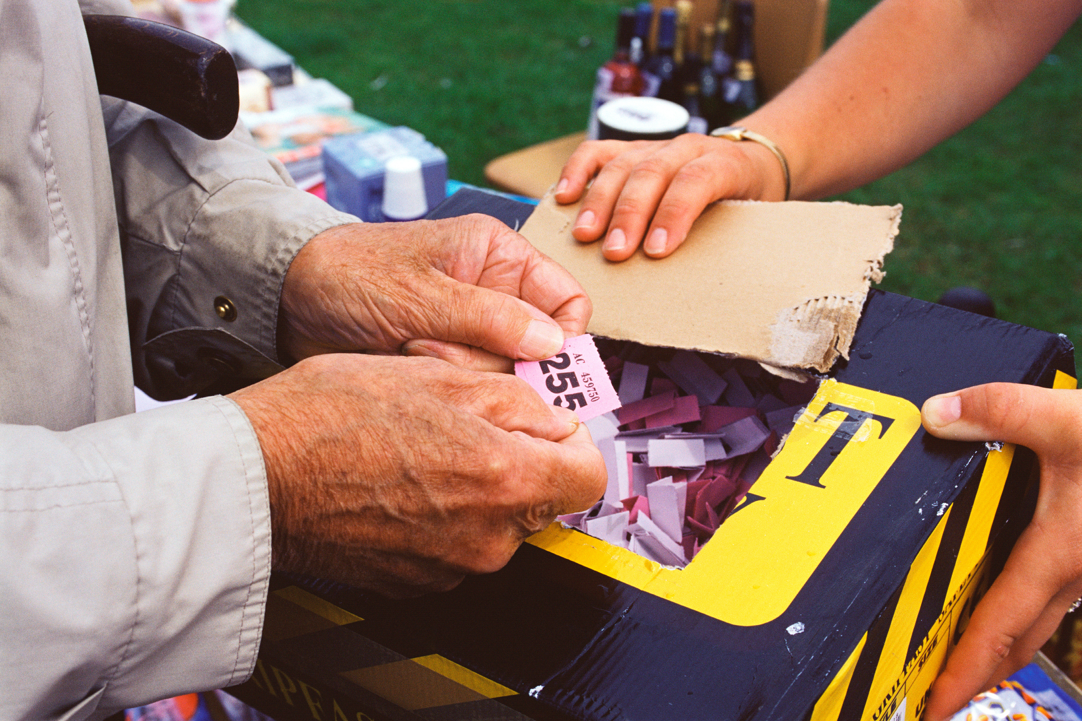 Two people draw raffle tickets from a box at an outdoor event. One person holds tickets while another reaches inside the box