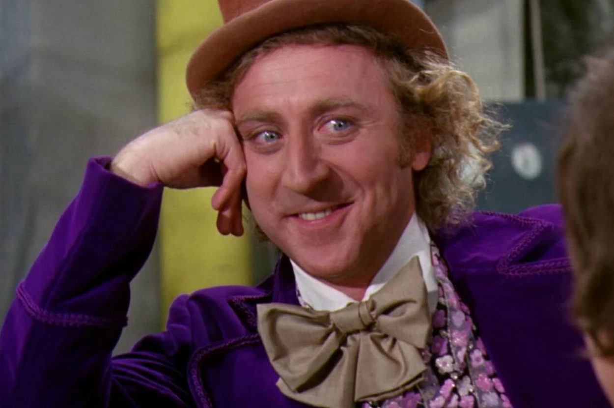 Gene Wilder, as Willy Wonka, smiles with his chin resting on his hand, wearing a top hat and a large bow tie with a purple suit and floral shirt