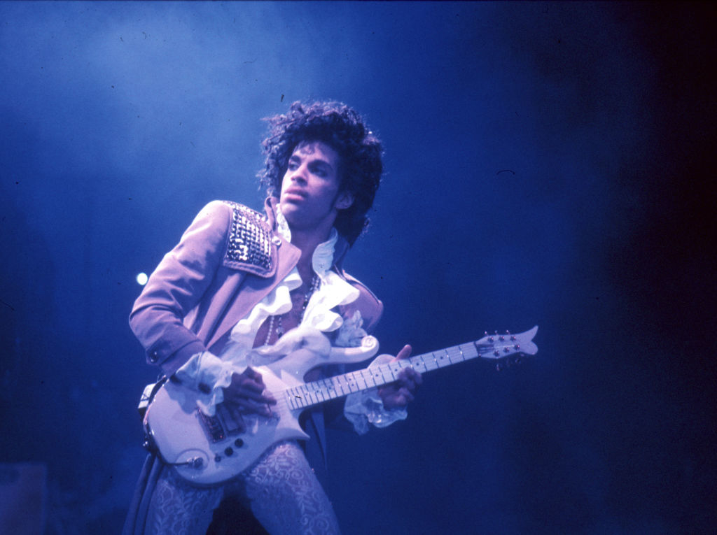 Prince playing a guitar on stage, wearing a ruffled shirt with a studded jacket, performing passionately