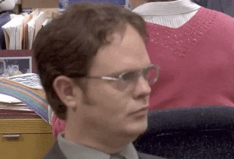 Dwight Schrute from The Office, wearing glasses and a suit, looks perplexed while seated at his desk in an office setting