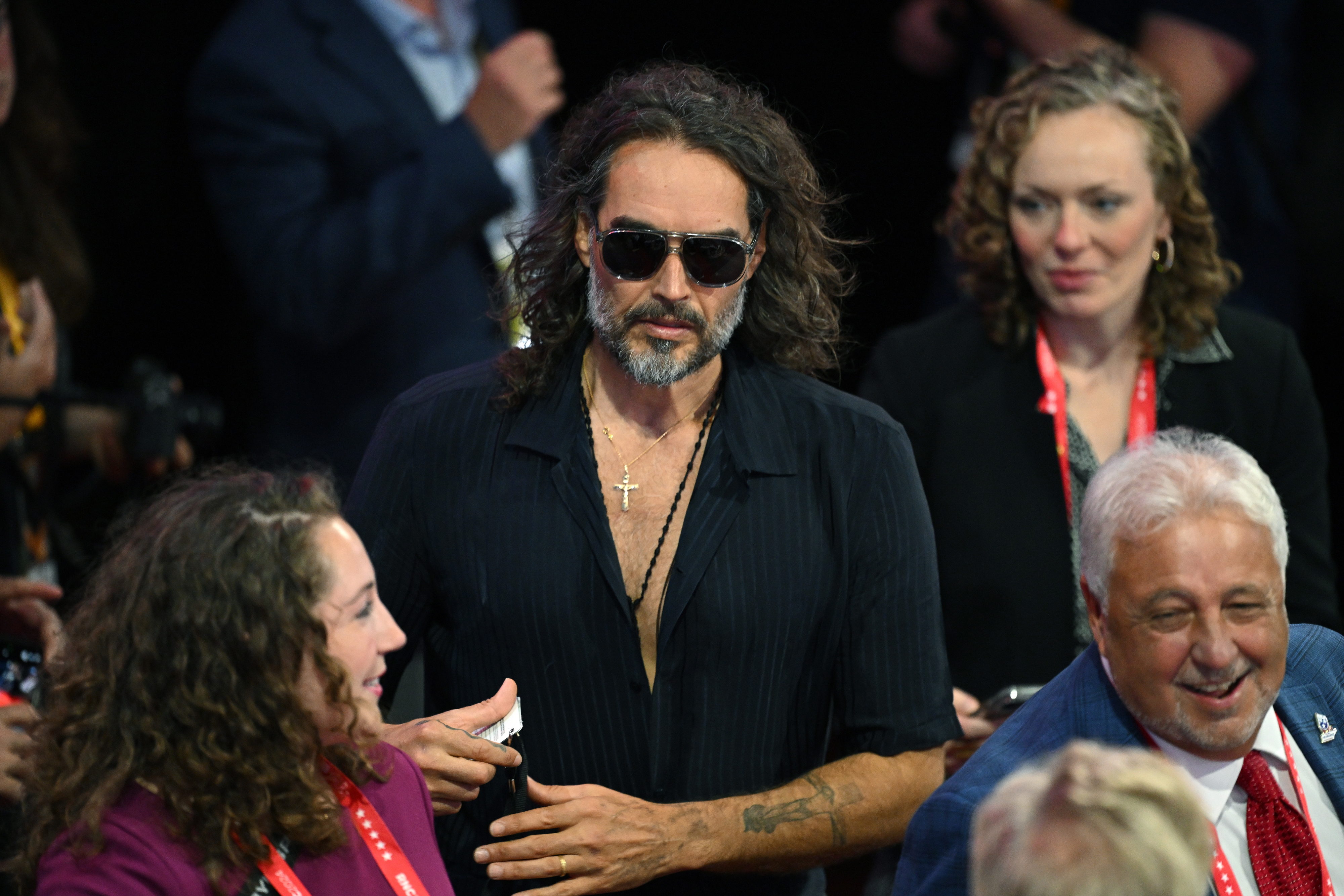 Russell Brand, with long hair, sunglasses, and a cross necklace, standing among a crowd. People around him are engaged in conversation