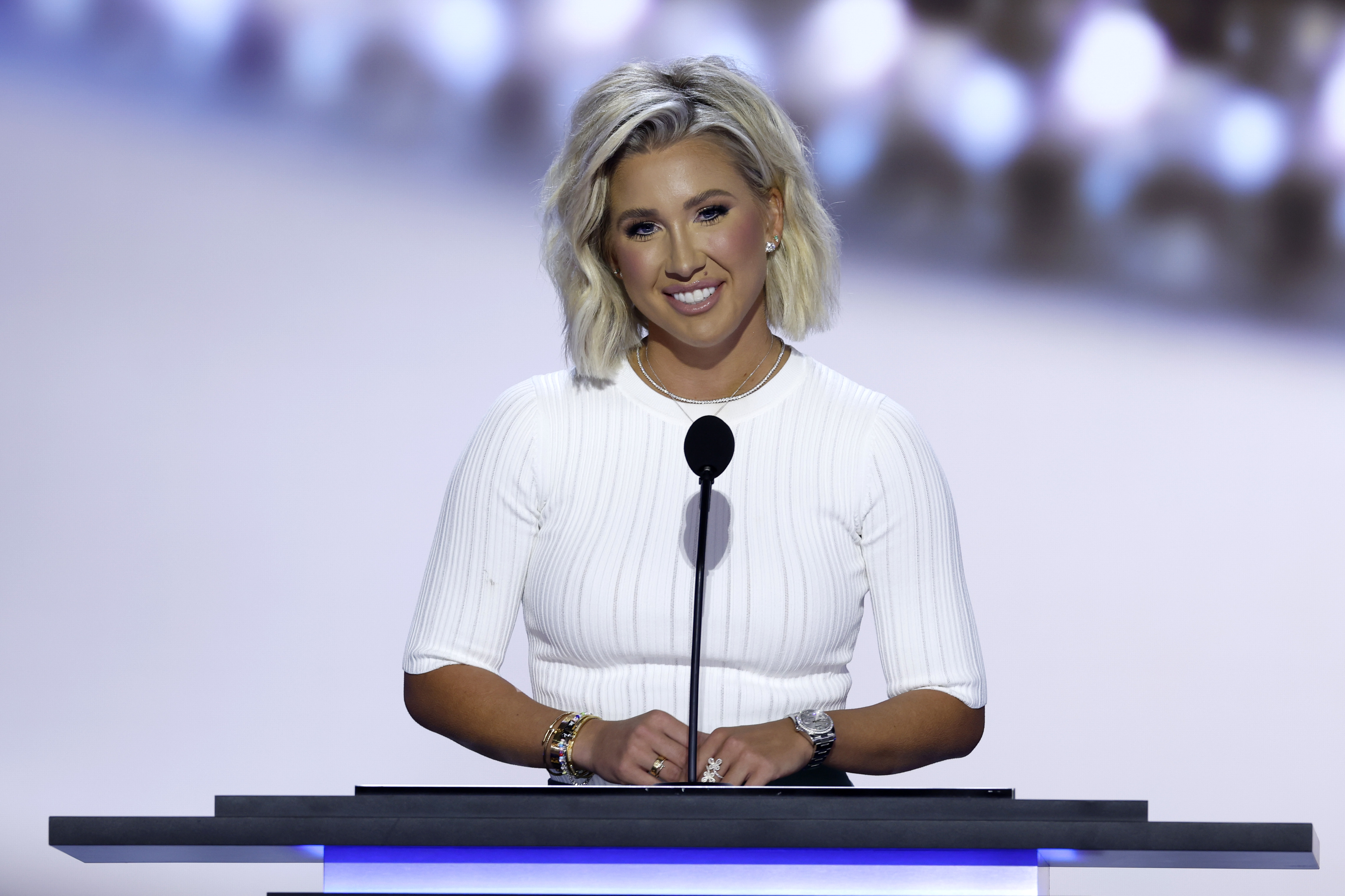 Savannah Chrisley, in a fitted white dress, stands behind a podium with a microphone, smiling during a speech or presentation