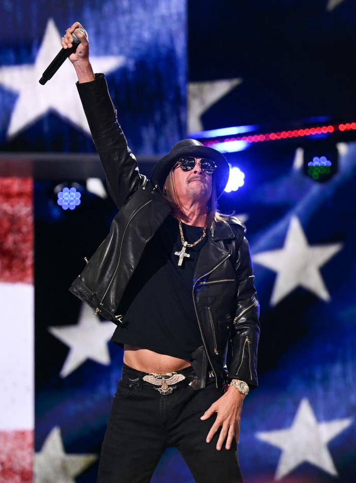 Kid Rock performs on stage wearing a black leather jacket, hat, sunglasses, and cross necklace, with an American flag backdrop
