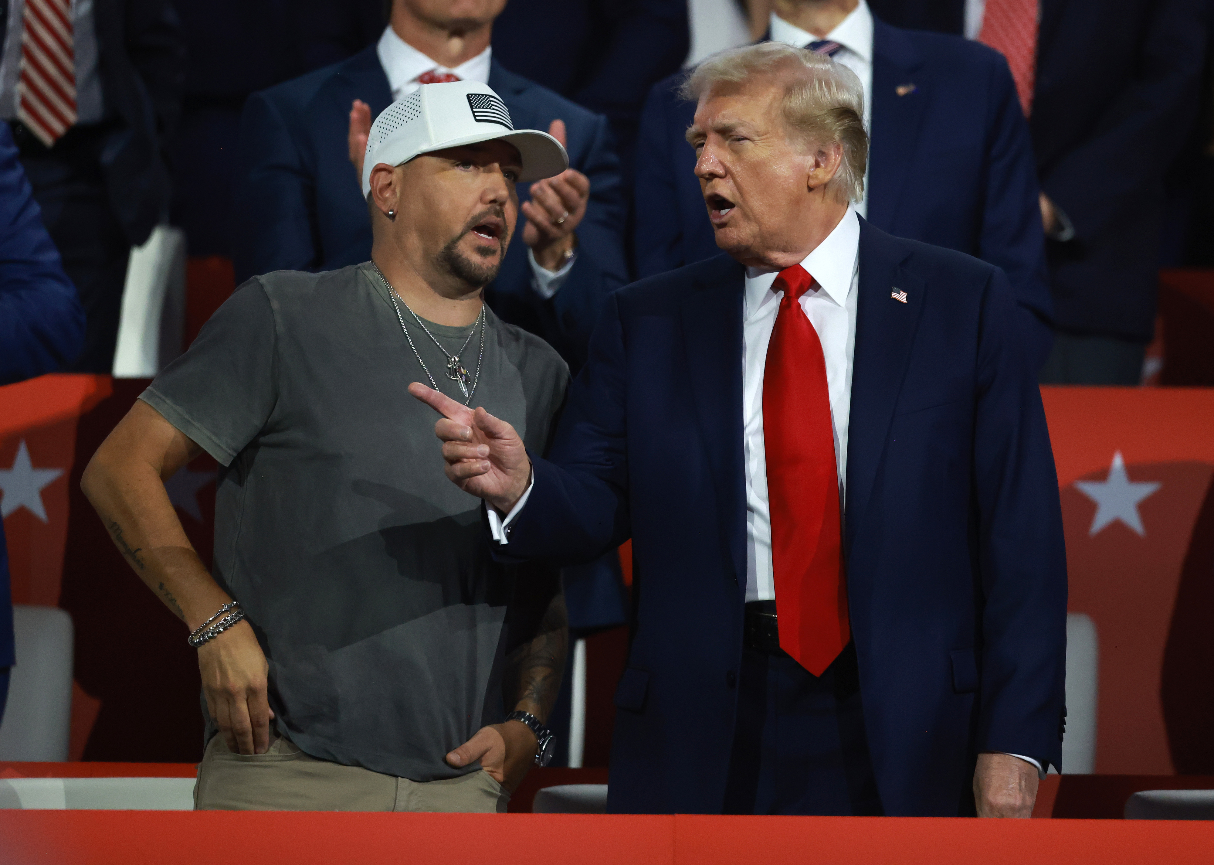 Jason Aldean and Donald Trump are talking animatedly at a public event, with Jason casually dressed in a t-shirt and cap and Trump in a suit